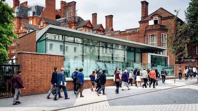 Group of people walking past a modern glass and brick building with historic red brick structures in the background.