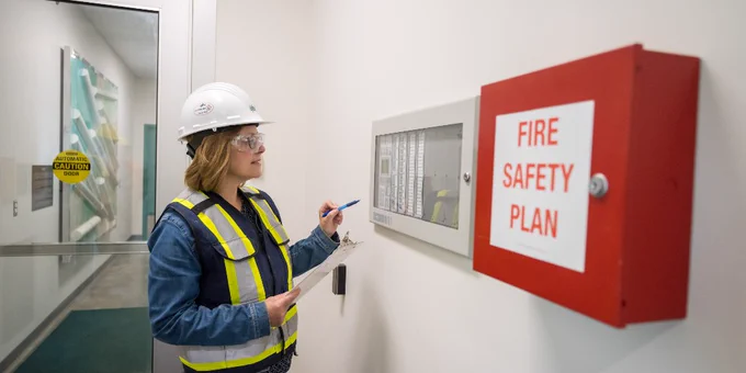 A person wearing a hard hat and safety vest is examining a control panel on a wall next to a red box labeled "Fire Safety Plan." The individual is holding a clipboard and pen. In the background, there is a doorway with a glass panel and a safety sign visible.