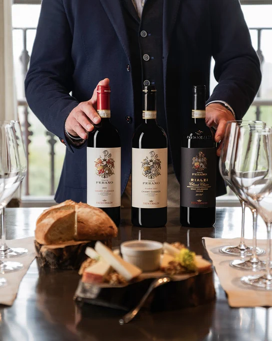A person arranging three Marchesi Frescobaldi wine bottles from Tenuta Perano in Chianti Classico on a table alongside bread and a wooden board.
