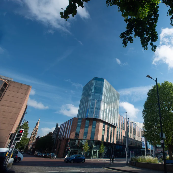 Modern glass and brick building under a bright blue sky with scattered clouds and leafy trees nearby.