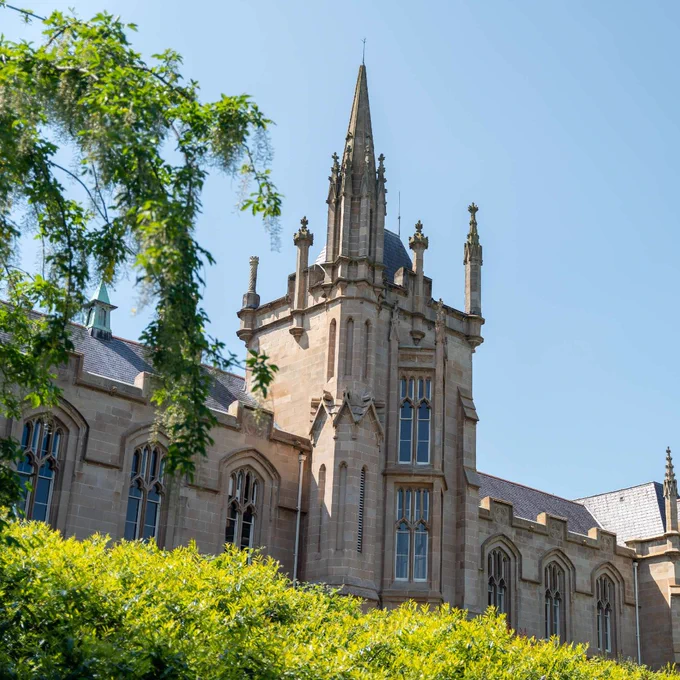 Gothic-style stone building with tall spires and arched windows under a clear blue sky, framed by green trees.