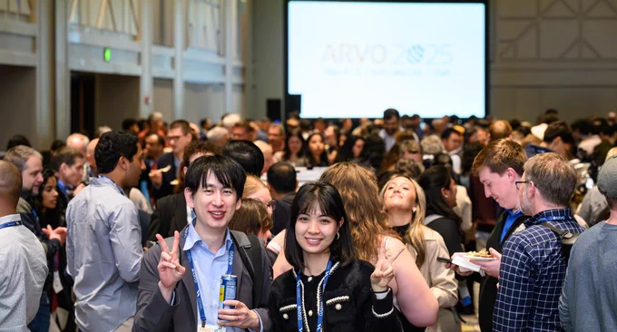 Social and professional setting with a large, diverse group of people gathered in a Convention Center hall, many looking towards the camera. Some are holding beverages and making peace signs. In the background, a large screen displays the ARVO Meeting logo.