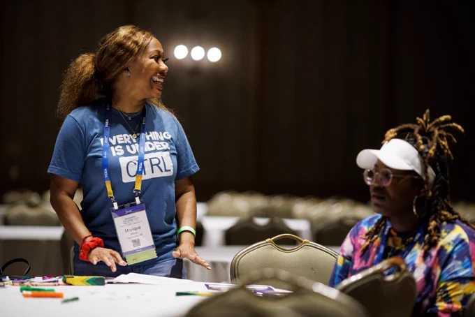 Photo of two people at a table. One person is sitting. The other person is standing up and smiling at something off camera. 