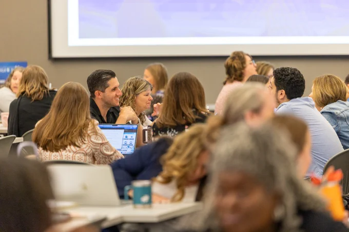 A group of ESC staff members is engaged in discussions at PD Day. Tables are filled with attendees, and a large screen is visible in the background.