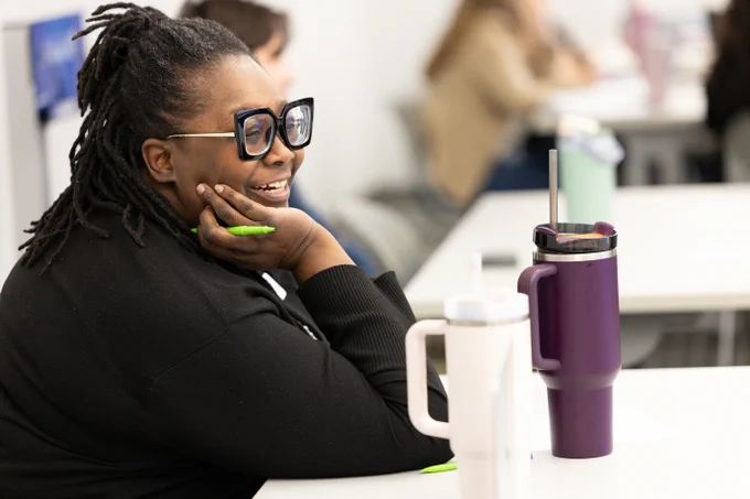 An ESC staff member wearing glasses is smiling and sitting at a table in a conference room. There are two large travel mugs on the table, one white and one purple. Other people are blurred in the background, seated at tables.