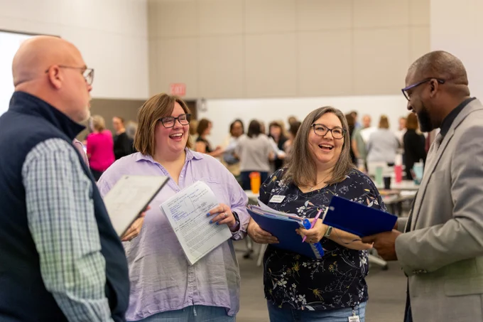 A group of ESC staff members is in a conference room, engaged in conversation and holding documents and notebooks. They are engaged in a lively discussion in a conference room with others in the background.