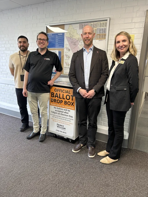 Commissioner Steven Koski and elections staff from Prince William County standing beside an official ballot drop box inside a building with a map on the wall behind them.