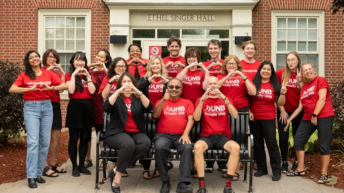 A group of people wearing red "UNB Proudly UNB" shirts and black pants stand and sit in front of a building labeled Ethel Singer Hall. Some individuals are making heart shapes with their hands. The group is smiling, and the setting has a friendly, collegiate atmosphere.