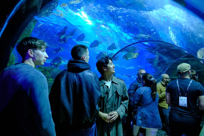 A group of people enjoy an aquarium scene, admiring fish swimming in vibrant blue water under a dome structure.