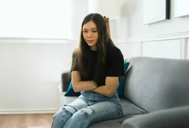 A young woman with long dark hair sits on a gray sofa with her arms crossed, looking down at a smartphone lying on the cushion next to her; she wears a black t-shirt and ripped blue jeans, appearing thoughtful or somber in a bright room with white walls and a large window in the background.