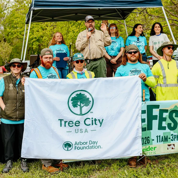 Group of volunteers holding a Tree City USA banner at an outdoor Arbor Day Foundation event under a canopy tent.