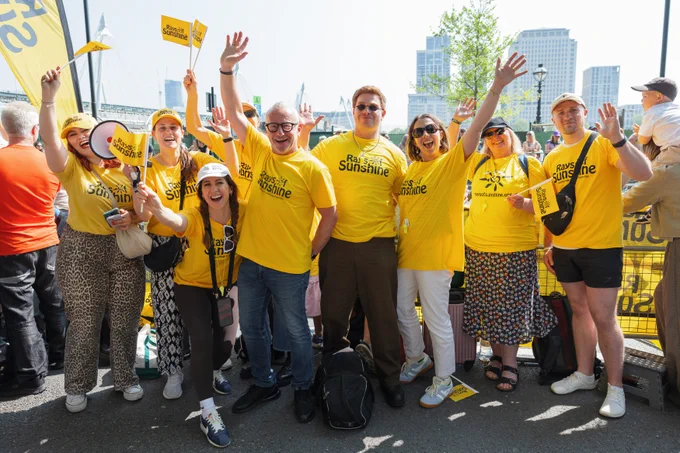 Smiling volunteers in yellow "Rays of Sunshine" shirts waving and holding flags at an outdoor charity event