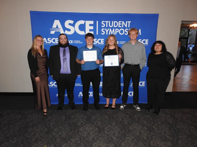 Timber Strong team stand in front of a blue ASCE Student Symposium backdrop, smiling and posing together, with two students holding award certificates after the competition.