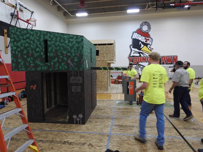 A nearly completed timber structure stands on a competition floor as team members move materials and equipment nearby, with another team’s structure visible in the background.
