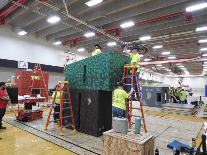 Students in bright yellow shirts and hard hats stand on ladders assembling a two-story timber structure with a green forest-themed exterior during a timed construction event inside a gymnasium.