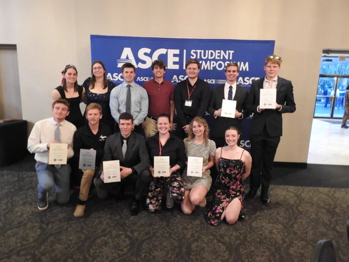 Steel Bridge team dressed-up students pose in front of an ASCE Student Symposium backdrop holding award plaques, smiling proudly after receiving competition honors.