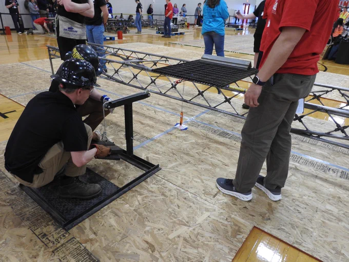 Students wearing hard hats carefully adjust a steel bridge structure during assembly on a competition floor, with one student crouched at a support frame while others observe and prepare for loading.