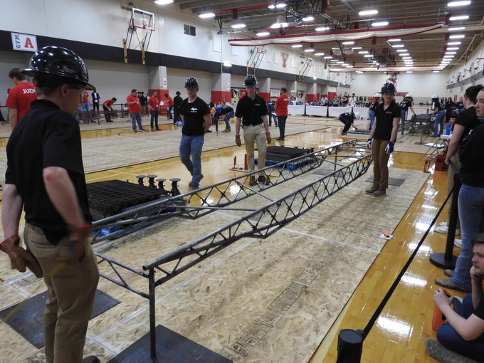 Multiple students rapidly place steel decking panels onto a partially assembled bridge during timed construction, demonstrating teamwork and speed in a gymnasium competition setting.