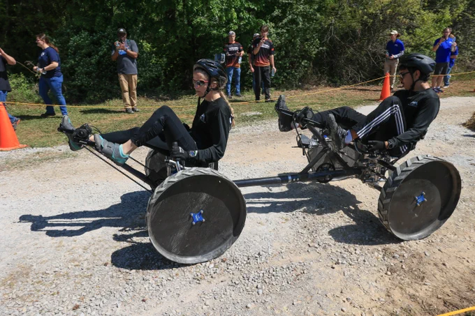 Two individuals wearing helmets race on custom-built, pedal-powered tricycles on a gravel path with spectators in the background.