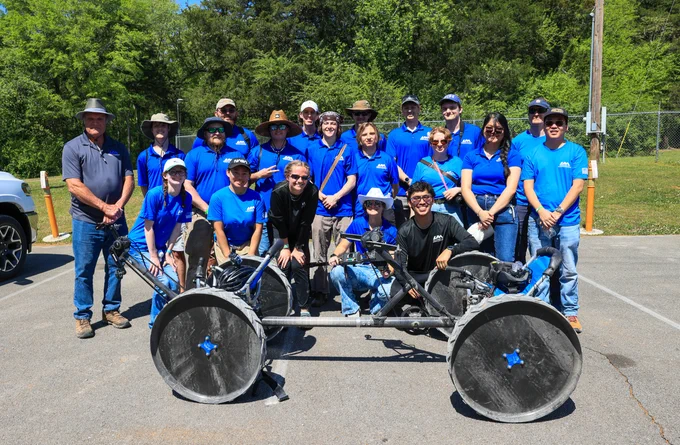 Group of UAH students in blue shirts posing outdoors behind their man-powered moonbuggy.