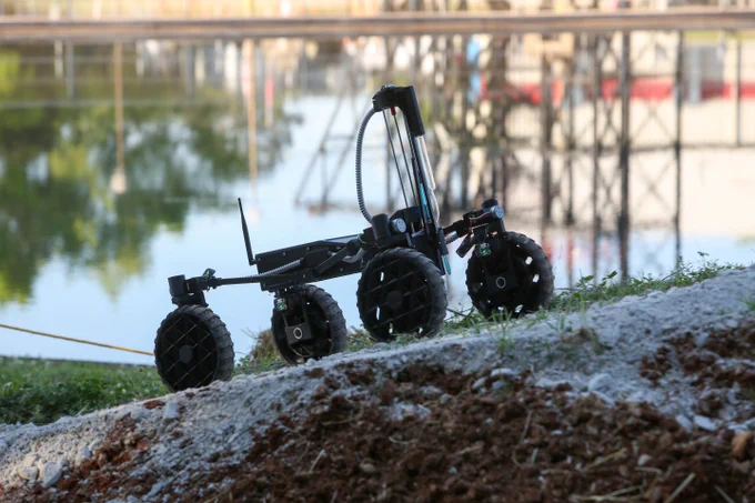 UAH's remote-controlled rover with large wheels positioned on a dirt mound near a reflective body of water and industrial structures.