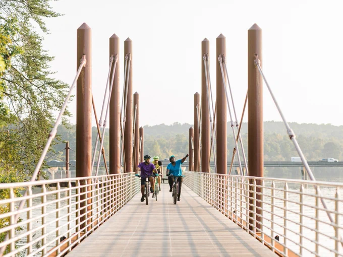 Three cyclists ride across a modern pedestrian bridge lined with tall metal posts and cables, with a river below, trees on the riverbank, and a distant highway visible in the background.