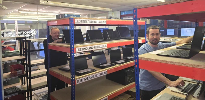 Two technicians working in the ST4G laptop testing room, filled with shelves labeled 'Testing and Installing' holding multiple laptops in various stages of setup.