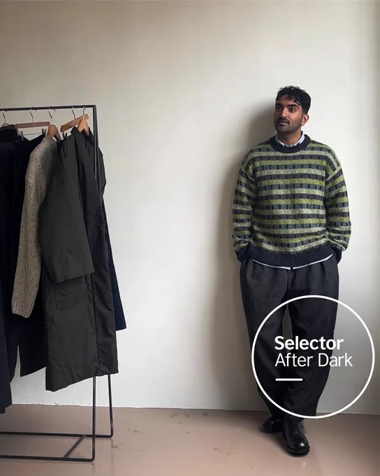 A man, Rohan Rakhit, is leaning casually against a blank wall in a striped knit jumper and wide-leg trousers, gazing to the right, with a clothing rail of muted coats beside him. The Selector After Dark logo sits in a white circle at the bottom right.