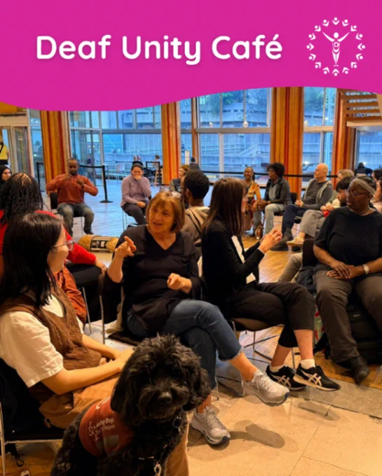 People seated in a circle signing and chatting at a Deaf Unity Café gathering, with a small service dog in the foreground.