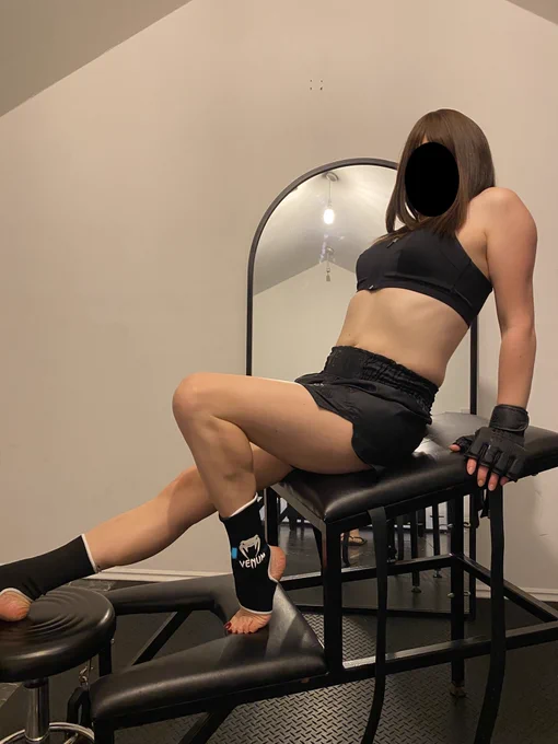 Trans Princess Jezebel poses on a rack, sitting with one leg splayed out and the other pulled back, arms extended behind her. She's wearing black kickboxing gear including Venum ankle bands, Muay Thai shorts, sports bra and sparring gloves.