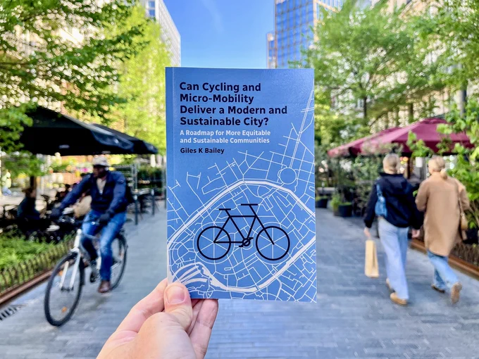 A hand holds a paperback book, "Can Cycling and Micro-Mobility Deliver a Modern and Sustainable City?" by Giles Bailey, in front of a pedestrianised street in the centre of Brussels.