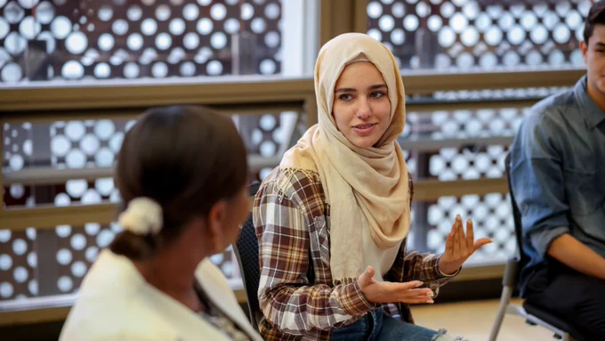 Support group. Two women chatting sitting on chairs, one women is in a hijab, one has dark hair in a ponytail.