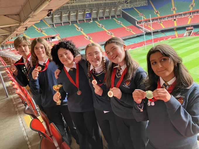 Six students in school uniform, some wearing medals and one holding a trophy, pose in stadium seating.