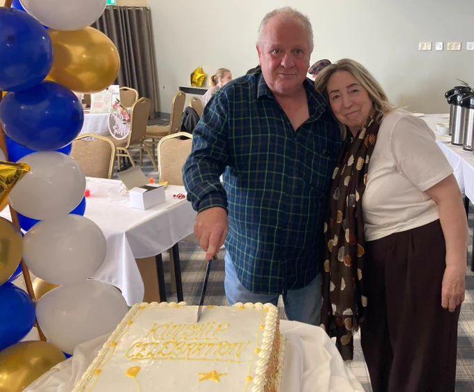 A man and woman cutting a cake that says "KINSHIP CELEBRATION" at a party with balloons and food.