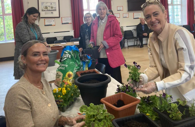 Women planting flowers around a table with compost and young plants in a community hall setting.