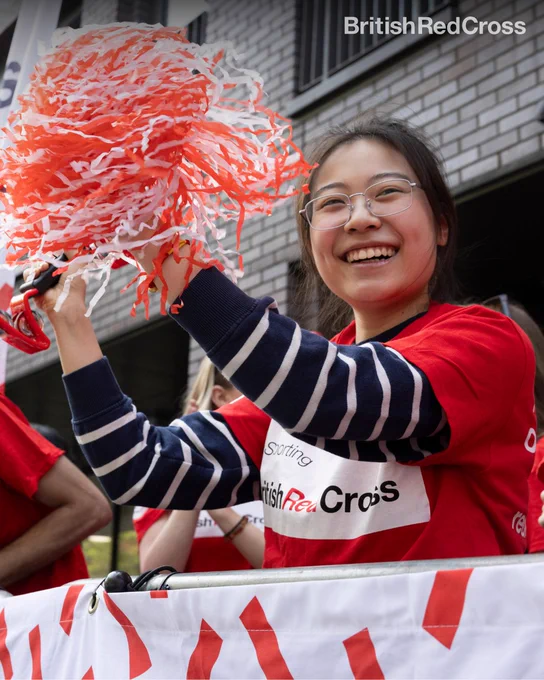 Image of Red Cross volunteer wearing a British Red Cross t-shirt holding up pom poms cheering from the sidelines. 
