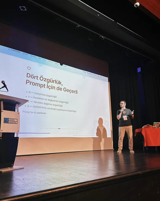 Bir konuşmacı sahnede mikrofonla sunum yapıyor. Ekranda “Dört Özgürlük, Prompt için de Geçerli” başlıklı bir slayt yer alıyor. Sahnenin solunda kürsü, sağında ise masa bulunuyor.