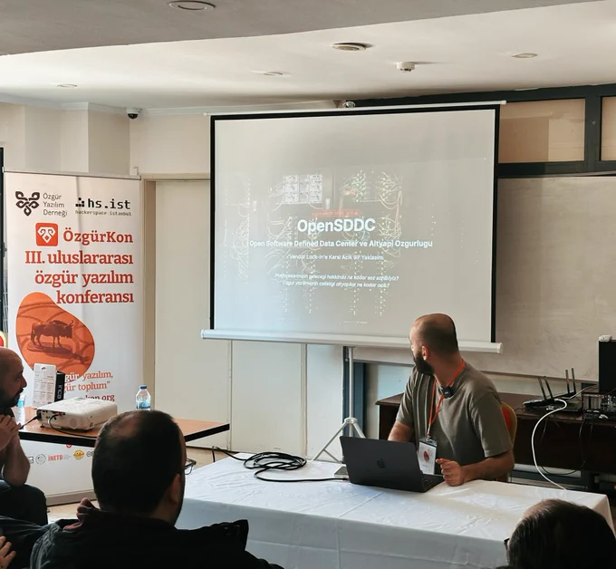 Bir atölye salonunda katılımcılar otururken, konuşmacı dizüstü bilgisayarıyla sunum yapıyor. Perdede “OpenSDDC” başlıklı bir sunum yer alıyor. Yan tarafta Özgür Yazılım Derneği ve ÖzgürKon bannerı bulunuyor.