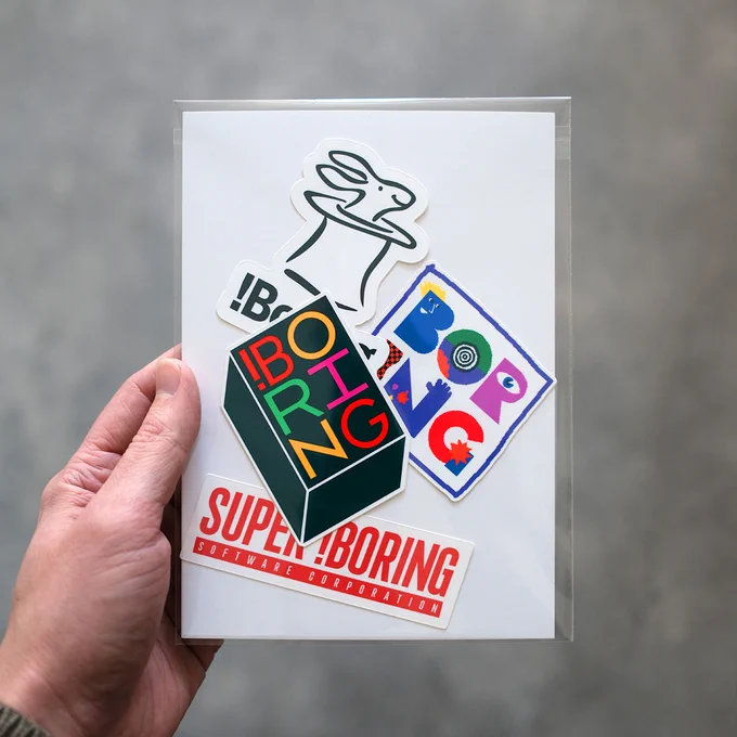 A hand holds a collection of colorful stickers featuring whimsical designs, including a rabbit, the word "BORING," and abstract shapes.