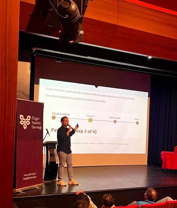 Bir konuşmacı sahnede mikrofonla sunum yapıyor. Ekranda web erişilebilirliği ve form tasarımıyla ilgili bir sunum slaytı yer alıyor. Ön planda dinleyiciler görülüyor.