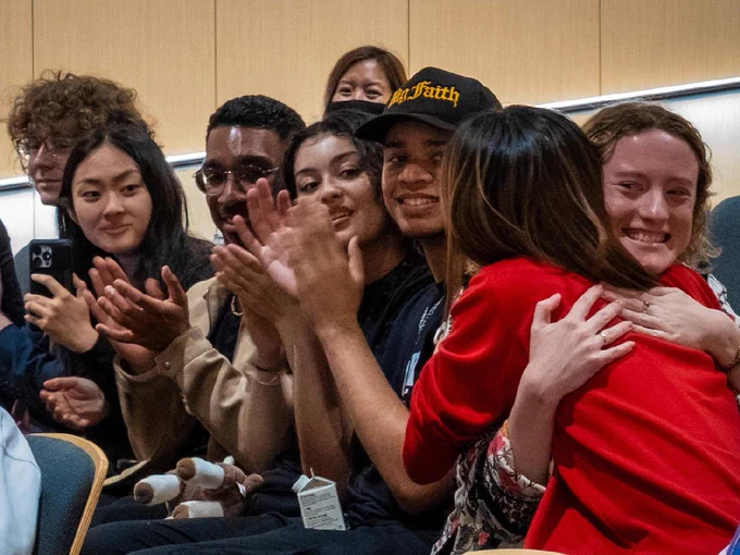 A group of people seated in an auditorium, some are smiling and clapping. One person wears a cap with "Faith" printed on it. Another person is being hugged by someone wearing a red garment. They appear engaged and cheerful.