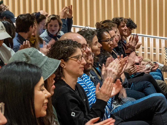 A diverse group of people seated in an auditorium are applauding. Everyone appears attentive and engaged. The background shows wooden paneling.
