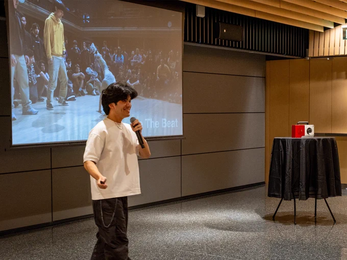 A person holds a microphone and is speaking in front of a large screen displaying an image of a dance battle scene. The screen shows several dancers and "The Beat" is visible. There is a table with a red object on top near the speaker. The room has a modern design with wood and metal accents.
