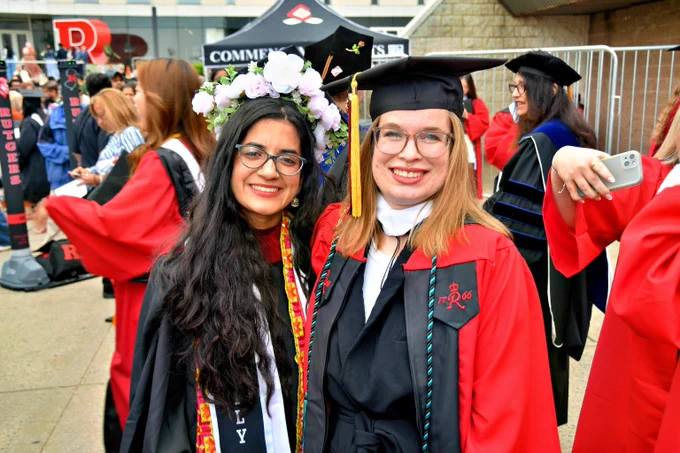 Two graduates in caps and gowns posing together, one wearing a flower crown and the other with a red academic robe and honor cords.
