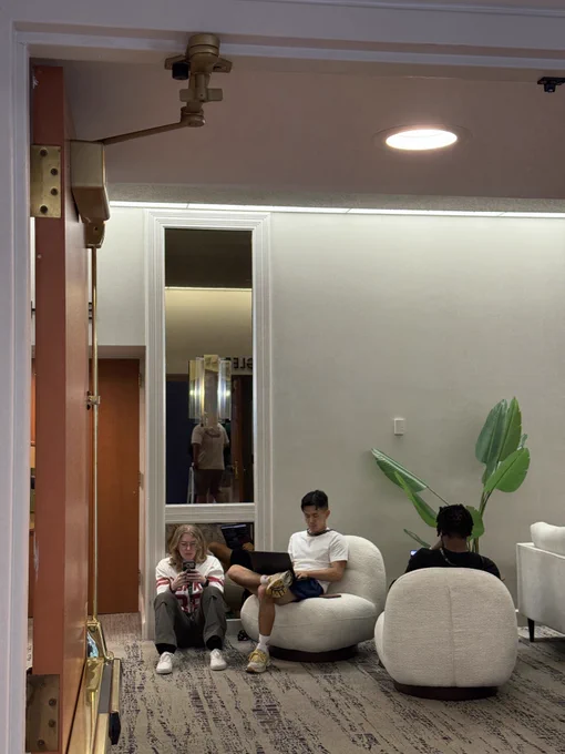 Photo taken during React Miami 2026 showing three people relaxing in a modern hotel or venue lobby. One person with long blonde hair sits on the floor against the wall, looking at their phone and wearing a graphic t-shirt and cargo pants. The photographer sits in a round, cream-colored boucle lounge chair wearing a white t-shirt and blue shorts, using a laptop. A third person in a black outfit sits in a matching boucle chair with their back to the camera, appearing to use a device. A large tropical bird-of-paradise plant stands in the corner against a light gray wall. The space features patterned carpet, recessed lighting, a tall brass floor lamp, and a mirror reflecting another attendee in the background. A security camera is visible mounted near the ceiling at the entrance.