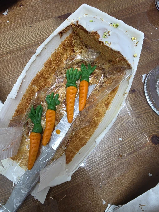 Das Bild zeigt die Überreste eines Rüeblikuchens (Karottenkuchens) in einer weißen Pappform, die auf einem Holztisch steht.
Es ist nur noch ein kleines Eckstück sowie Krümel am Boden der Form übrig. Der Kuchen ist mit einer weißen Zuckerglasur überzogen und mit gehackten Pistazien bestreut.
In der Mitte der Form liegen vier  Marzipan-Rüebli (Möhren) auf einer transparenten Plastikfolie.