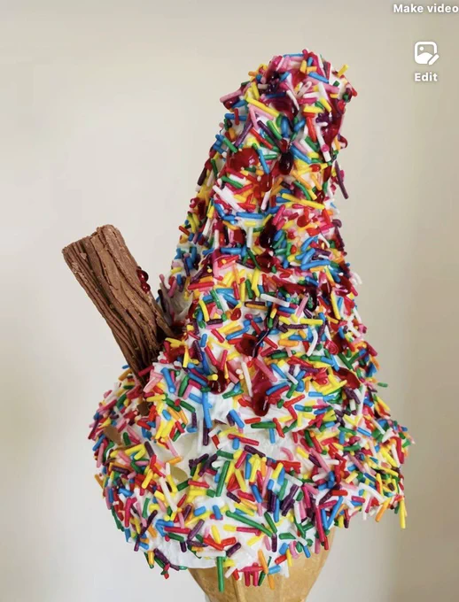 A close-up photograph of a tall, soft-serve vanilla ice cream cone. The ice cream is heavily coated in vibrant, multi-colored sprinkles (hundreds and thousands) and drizzled with bright red strawberry syrup. A chocolate flake bar is tucked into the side of the swirls. The cone is made of a light golden waffle-style wafer, and the entire treat is set against a plain, neutral-toned background that makes the colorful toppings stand out.