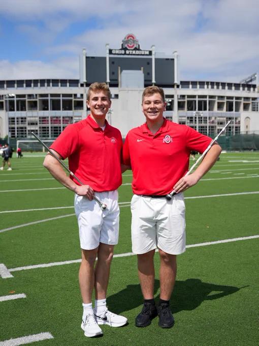 Ben Fox (left) and Jacob Snyder (right) are standing on the football field outside of  Ohio Stadium, both wearing red shirts and white shorts, holding batons.