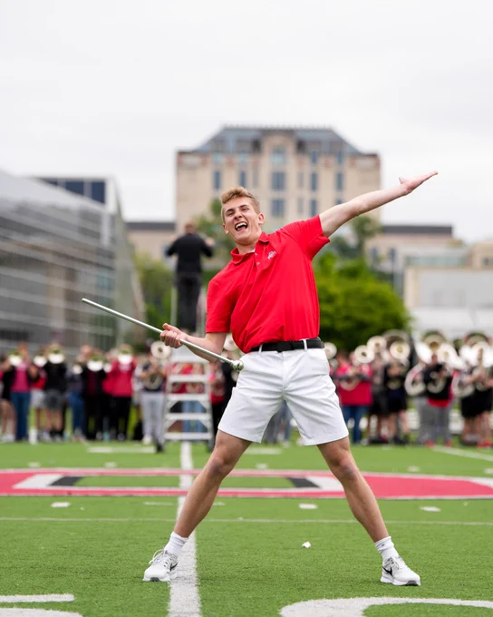 Ben, who is in a red shirt and white shorts energetically poses with a baton on a football field, with a marching band in the background. Buildings and trees are visible in the distance.