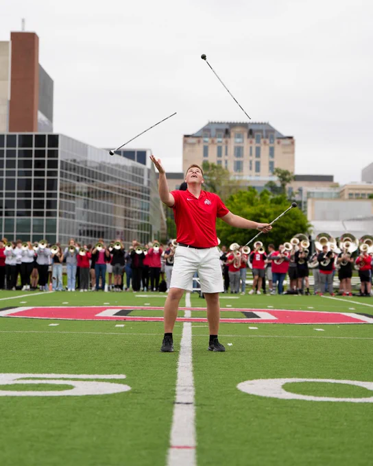 Jacob is in a red shirt and white shorts and is twirling three batons. A band is in the background, with buildings visible on a cloudy day.